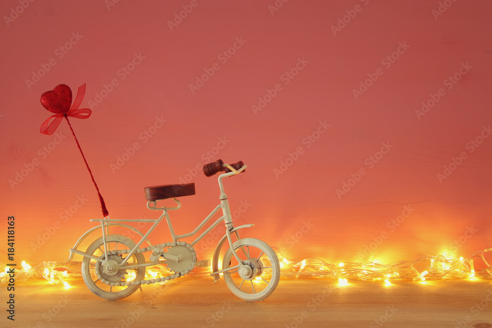 Fototapeta premium Valentines day romantic background with white vintage bicycle toy and glitter red heart on it over wooden table.
