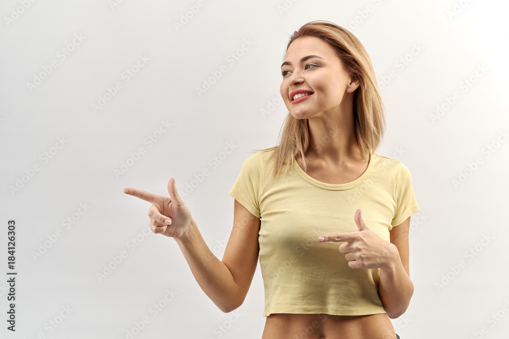 smiling young woman girl in a short light yellow t-shirt looking to the side and points his index fingers at an object. Joyful smile, positive emotions