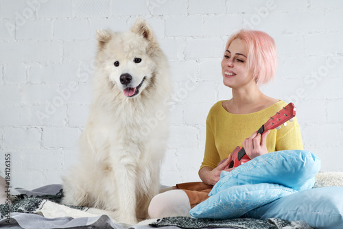 young friendly joyful girl playing the ukulele, the guitar sitting on the couch with her friend a big white dog.