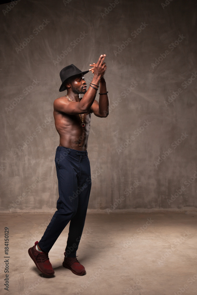 Fit strong physique African American young man wearing black hat and trousers posing dancing near gray concrete cement wall in studio. Fit strong physique African American young man wearing black hat and trousers posing dancing near gray concrete cement wall in studio.