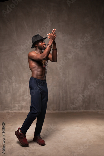 Fit strong physique African American young man wearing black hat and trousers posing dancing near gray concrete cement wall in studio.