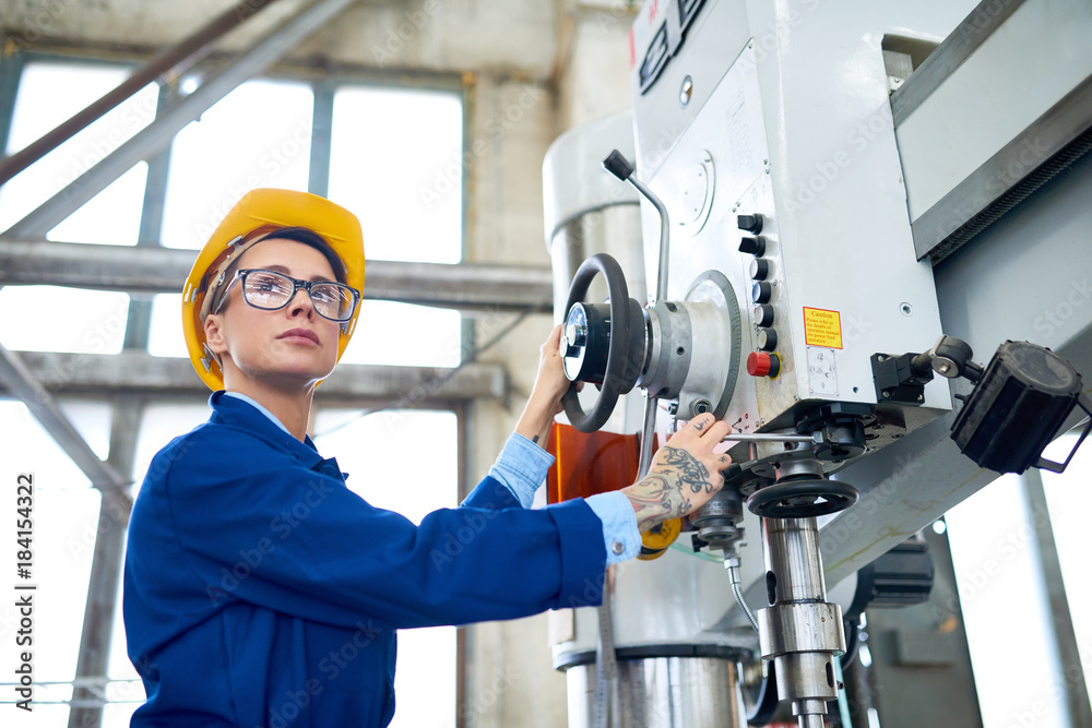 Portrait of attractive plant worker wearing eyeglasses and protective helmet operating bench drilling machine at spacious production department