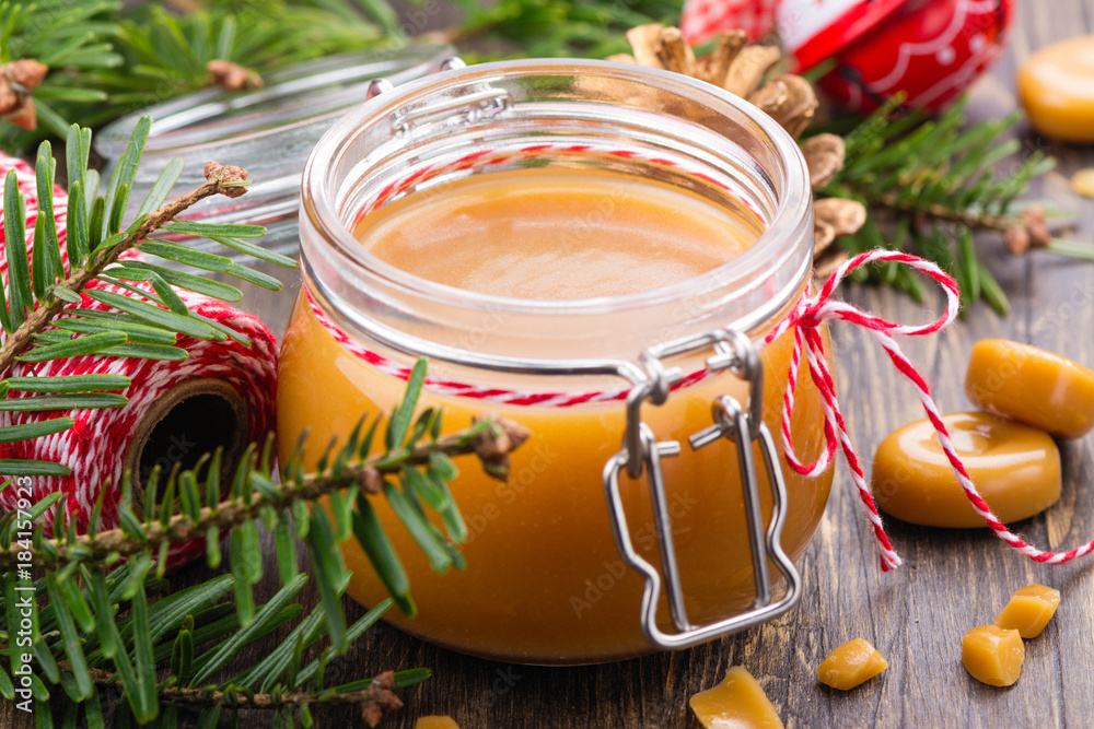 Homemade salted caramel sauce in a glass jar and Christmas decor on wooden background Homemade salted caramel sauce in a glass jar and Christmas decor on wooden background