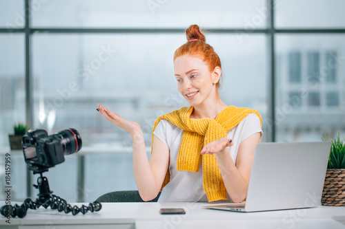 Young woman recording video on camera mounted on tripod for her vlog. Pretty woman smiling at camera sitting at table with laptop photo