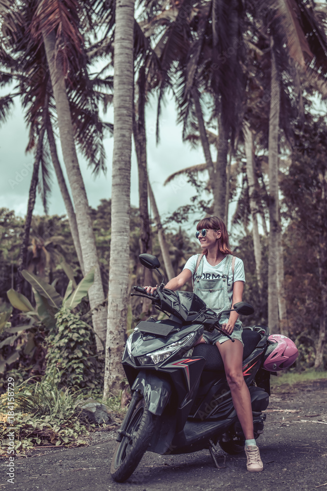 Young happy woman on a motorbike in the jungle rainforest of a tropical Bali island, Indonesia. Freedom concept. Lady on a scooter.