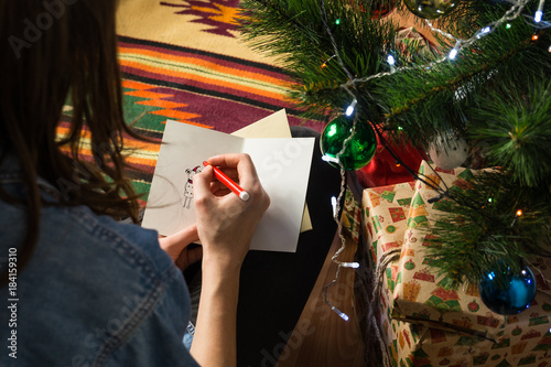 Young female signs postcard sitting on floor next to decorated fur tree and pile of new year presents packaged in beautiful packpaper photo