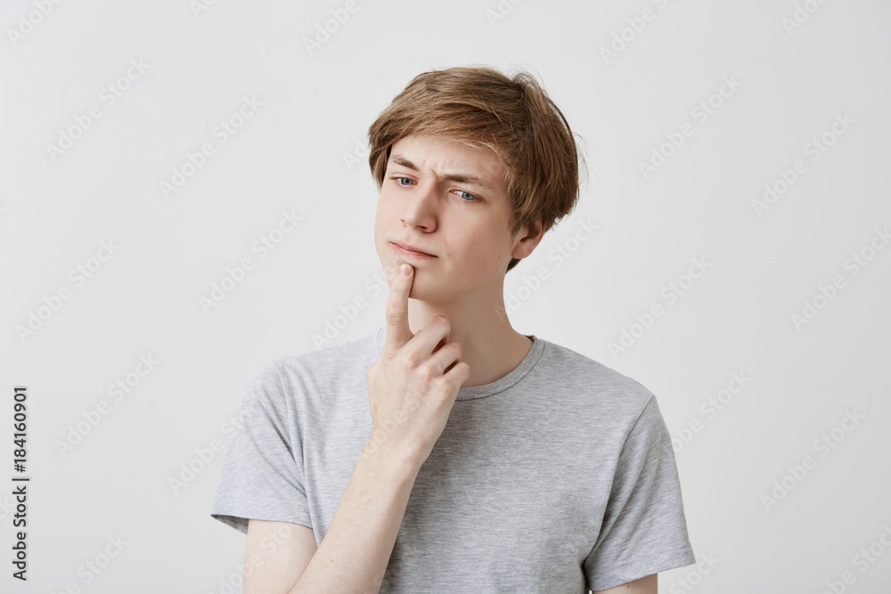 Attractive young fair-haired man with blue eyes dressed casually has thoughtful expression, keeps index finger on his chin, tries to recollect something or has great idea. Pensive male student