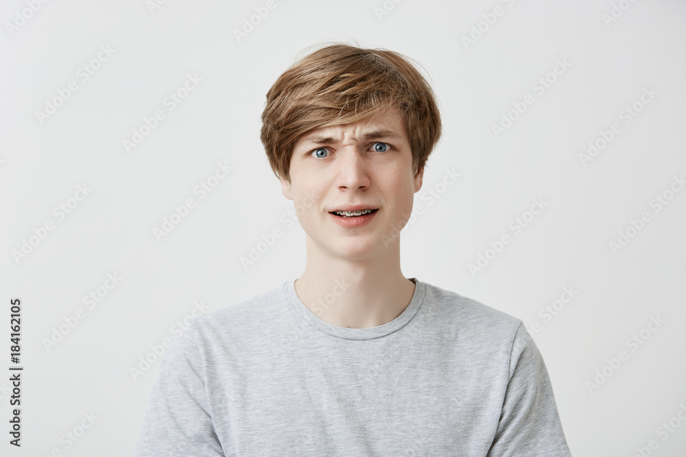 Indoor portrait of serious dissatisfied puzzled emotional fair-haired young man with braces, frowns his face in bewilderment, reacts on teachers`s remark. Peope and reaction concept Indoor portrait of serious dissatisfied puzzled emotional fair-haired young man with braces, frowns his face in bewilderment, reacts on teachers`s remark. Peope and reaction concept
