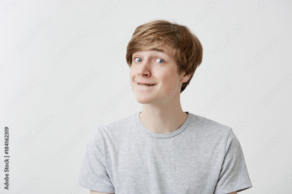 Good-looking male model with braces presses lips, looks pensively aside, thinks over his future actions or plans, wears light gray t-shirt, has clever expression, isolated against gray background Good-looking male model with braces presses lips, looks pensively aside, thinks over his future actions or plans, wears light gray t-shirt, has clever expression, isolated against gray background
