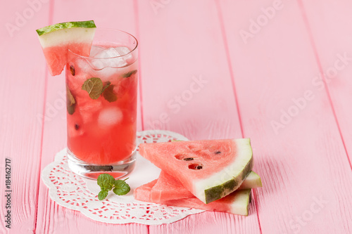 Watermelon lemonade with ice and mint leaves. Homemade lemonade of ripe berry with red and green ripes. Glass of cold watermelon tea. Refreshing summer drink. Cocktail on a wooden background