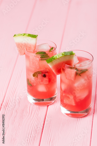 Watermelon lemonade with ice and mint leaves. Homemade lemonade of ripe berry with red and green ripes. Glass of cold watermelon tea. Refreshing summer drink. Cocktail on a wooden background