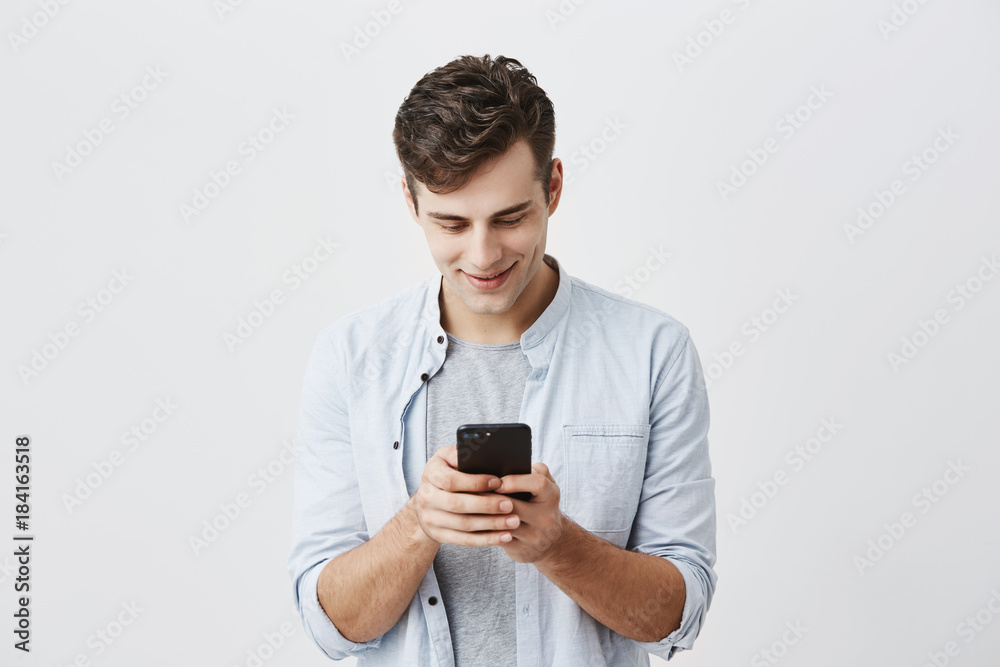 Handsome joyful student with dark hair wearing blue shirt messaging, typing message, using free onlipe app on his smartphone, looking at screen with smile, posing against gray background. Handsome joyful student with dark hair wearing blue shirt messaging, typing message, using free onlipe app on his smartphone, looking at screen with smile, posing against gray background.