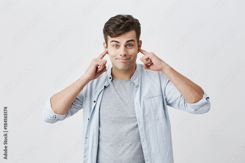 Portrait of angry and frustrated handsome man in blue shirt stopping his ears, plugging them with fingers, pursing lips while suffering from loud noise. Negative emotions. Stop this sound! Portrait of angry and frustrated handsome man in blue shirt stopping his ears, plugging them with fingers, pursing lips while suffering from loud noise. Negative emotions. Stop this sound!
