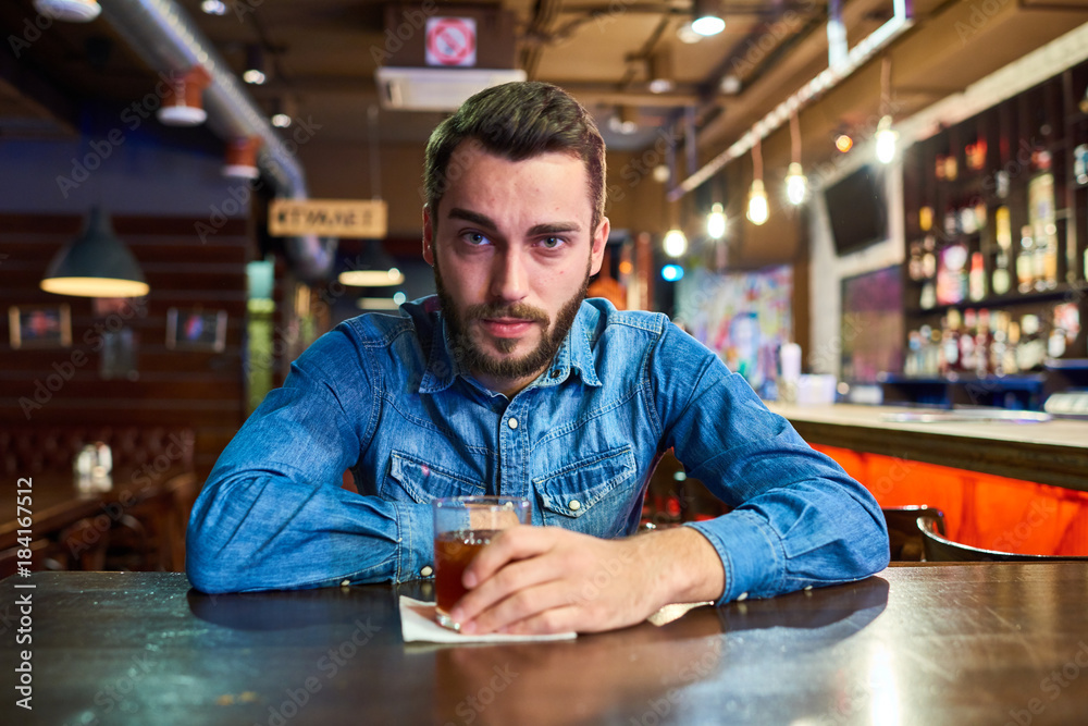 Portrait of drunk young man looking at camera with red puffy eyes sitting at table in pub with glass of alcohol Portrait of drunk young man looking at camera with red puffy eyes sitting at table in pub with glass of alcohol