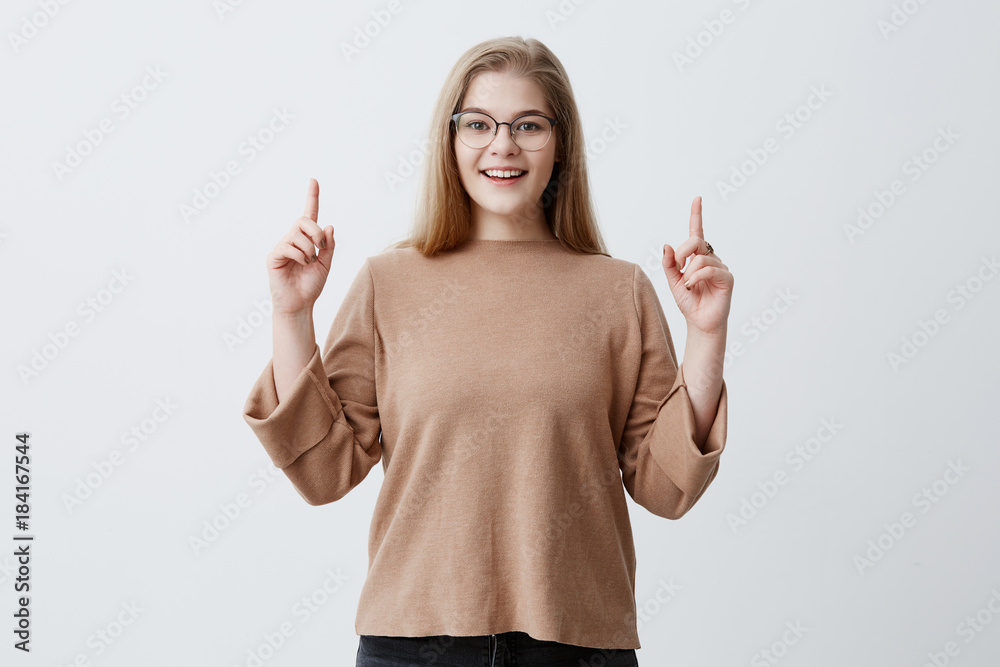 Advertisment concept. Happy woman in sweater, points up with fore fingers as shows something, has surprised pleased expression. Blonde female wearing stylish eyewear gestures against gray wall Advertisment concept. Happy woman in sweater, points up with fore fingers as shows something, has surprised pleased expression. Blonde female wearing stylish eyewear gestures against gray wall