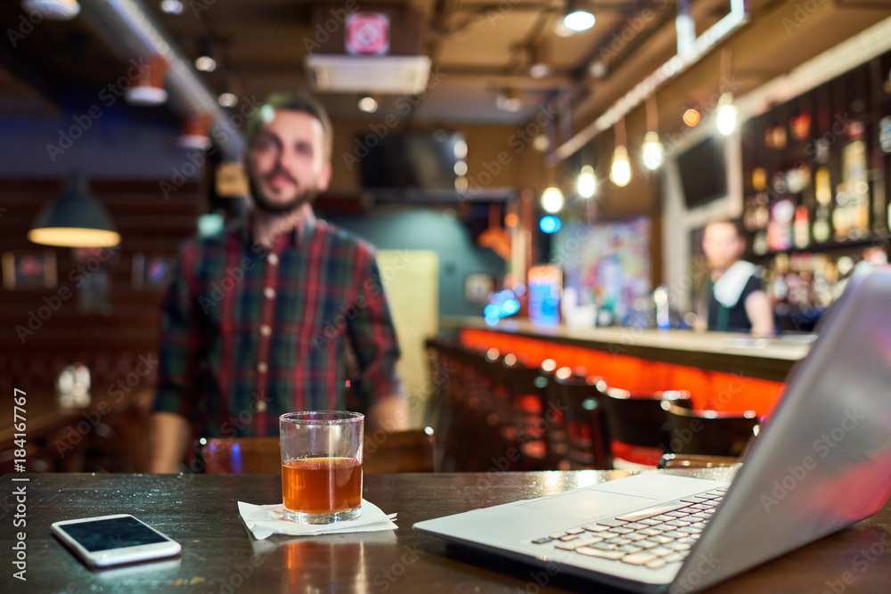 Laptop and glass of whiskey on bar counter in foreground with blurred figure of modern bearded man behind it, copy space
