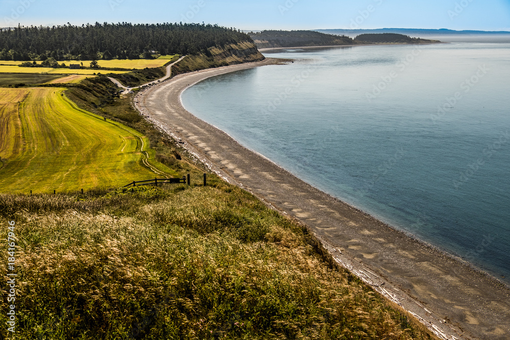 Curved stretch of beach between water and a bluff with islands and fog in the distance.