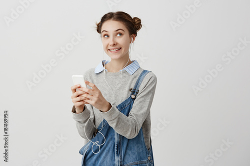 Portrait of attractive childish woman in denim jumpsuit looking sideways with joyful emotions. Woman in love receiving pleasant message on her smartphone feeling happiness. Facial expressions