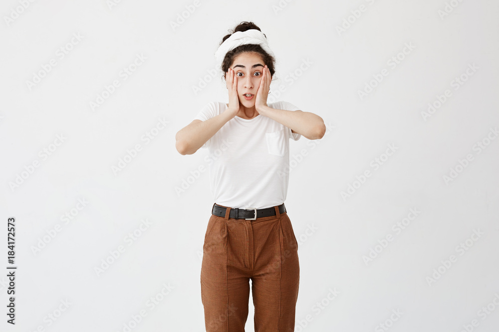 Indoor shot of scared dark-haired female with hairbun looks with bugged eyes and opened mouth, frightened to see awful things, can`t believe her eyes. People, emotions, feelings concept Indoor shot of scared dark-haired female with hairbun looks with bugged eyes and opened mouth, frightened to see awful things, can`t believe her eyes. People, emotions, feelings concept