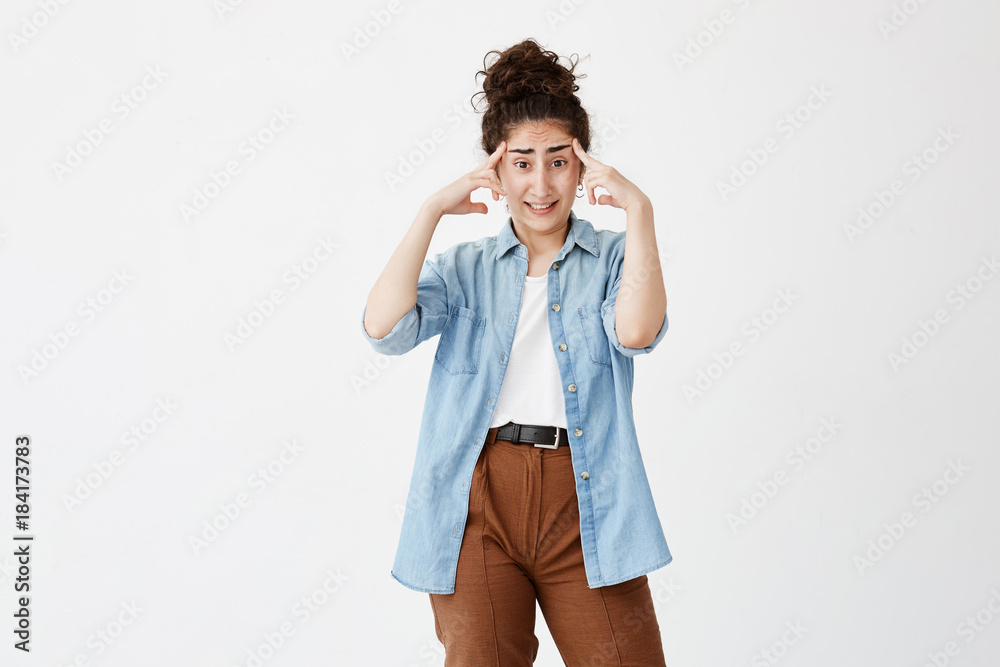 Brunette female with hair bun has headache after noisy party, clenches teeth and holds index fingers on temples. Tired young female worker overworked after writing advertising content Brunette female with hair bun has headache after noisy party, clenches teeth and holds index fingers on temples. Tired young female worker overworked after writing advertising content