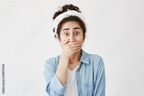 Shocked scared young brunette wearing do-rag and denim shirt covering mouth with hand keeping a secret. Dark-haired young female doesn't want to spread rumors, poses with popped eyes photo