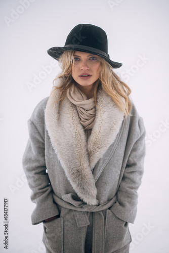 Portrait of beautiful blonde woman in hat and grey coat with snowy background