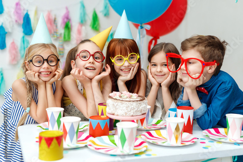 Portrait of funny group of children wear party caps, big spectacles, look with big appetite on birthday cake, want to taste it, have special occasion, spend time happily. Celebration, holidays concept photo