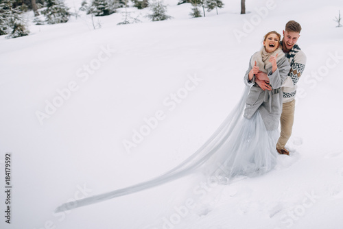 Beautiful couple stands in winter pine forest, woman in grey wedding dress and long veil, bearded man in sweater © Vadim Pastuh