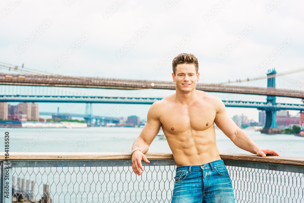 Man's Beauty in New York. Shirtless, half naked, waring jeans, a young,  strong, sexy guy casually standing at harbor by East River, smiling,  looking at you. Manhattan, Brooklyn bridges on background.. Stock