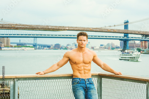 Man’s Beauty in New York. Shirtless, half naked, waring jeans, a young, strong, sexy guy casually standing at harbor by East River, looking at you. Manhattan, Brooklyn bridges, boat on background..