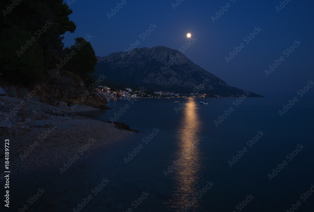 Night Picture of city Gradac in Croatia placed on the dalmatian coast in Makarska riviéra. Picture shown moon light in the water, pebble beach, mountain above the city and city lights.