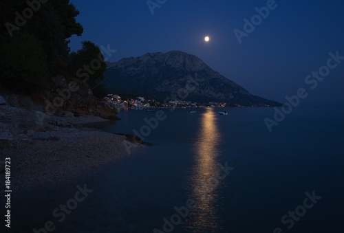 Night Picture of city Gradac in Croatia placed on the dalmatian coast in Makarska riviéra. Picture shown moon light in the water, pebble beach, mountain above the city and city lights.