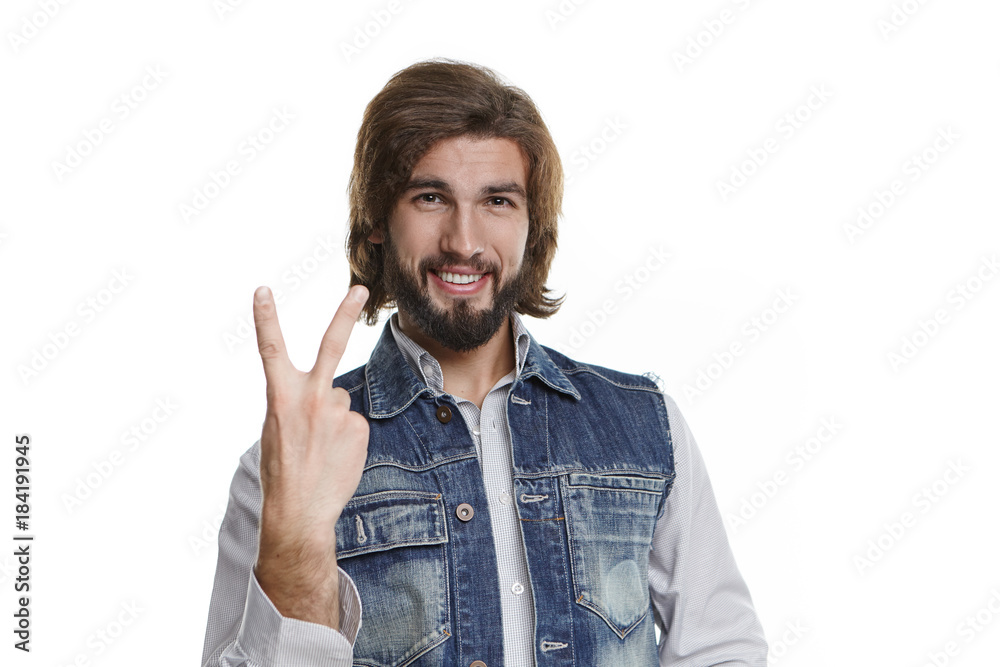 Horizontal shot of cool friendly looking hipster guy with thick beard and voluminous hair grinning broadly and showing peace or victory sign, expressing his positiveness, excitement and joy