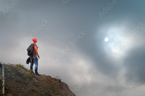 Sporty guy with a backpack on the edge of the cliff against a stormy clouds and sun. Travel concept.