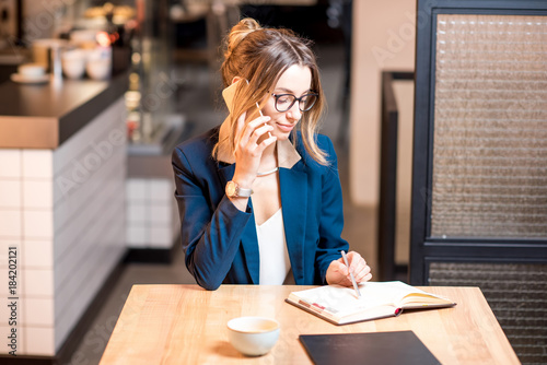 Young businesswoman strictly dressed in suit talking with phone working at the modern cafe interior