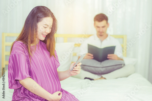 Asian pregnant people and expectation concept - happy pregnant woman holding mobile phone sitting on bed and touching belly at home with father reading book background.