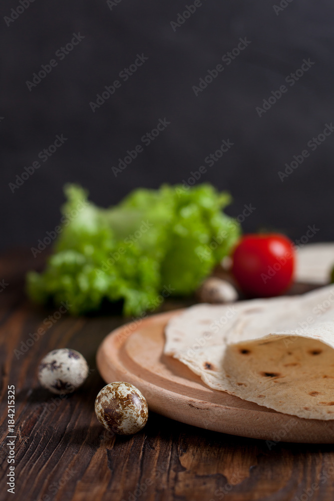 cooking burrito with greens, cucumbers, tomatoes and sausages on a round wooden board on a wooden background