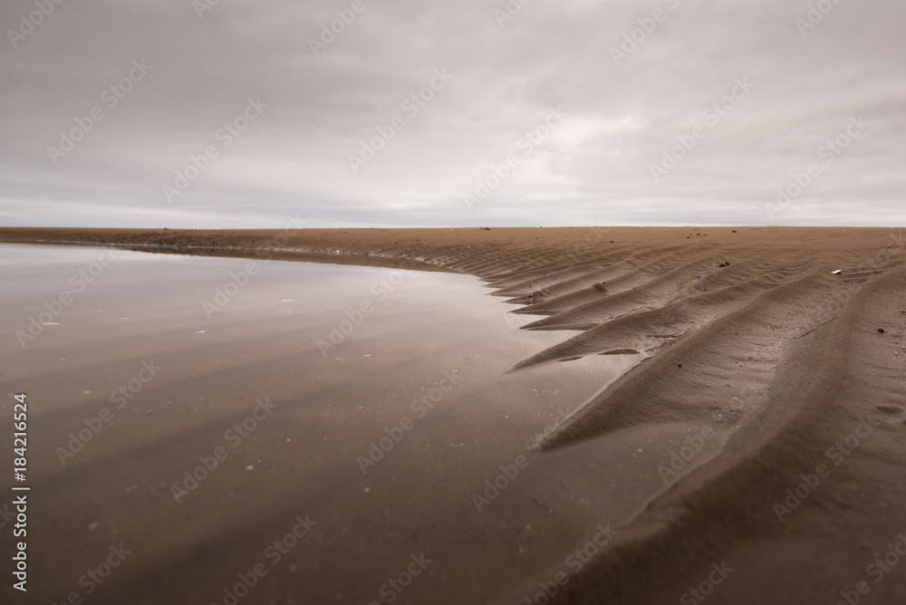 Sand ridges disappear into the water at the edge of a sandbar with a cloudy sky.