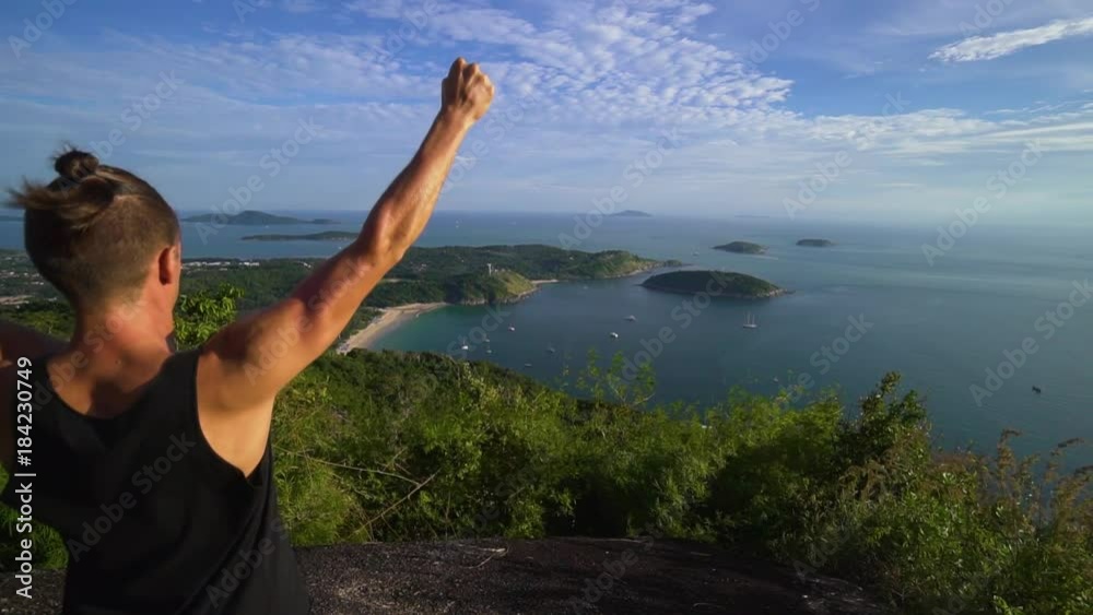 Athletic young man stands with his hands up on the edge of the mountain overlooking the sea and the islands. Celebrates the victory. Rear back view. Phuket. Thailand. Athletic young man stands with his hands up on the edge of the mountain overlooking the sea and the islands. Celebrates the victory. Rear back view. Phuket. Thailand.