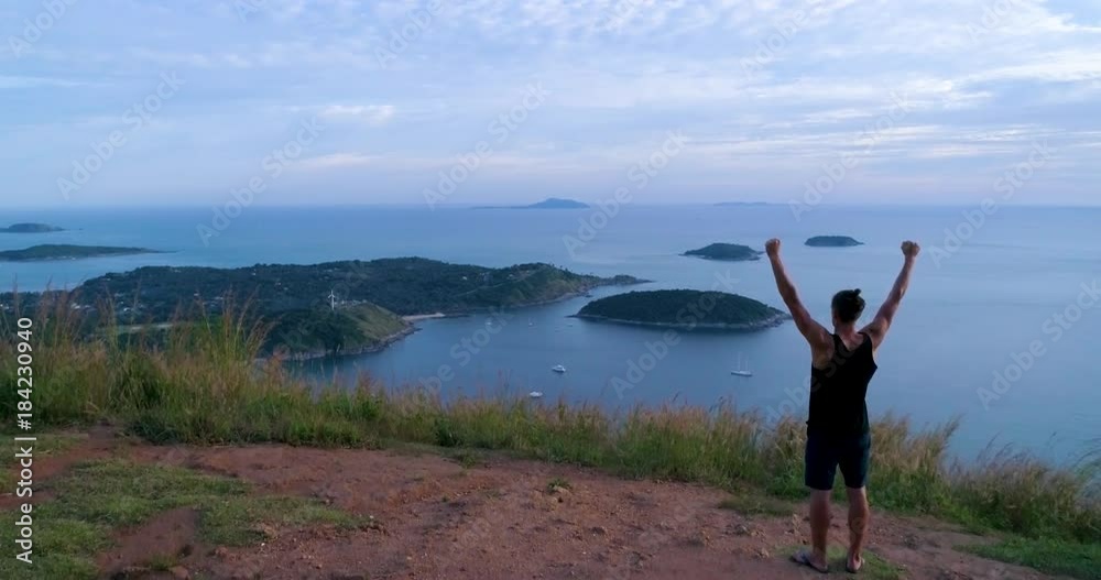 Aerial: Athletic young man stands with his hands up on the edge of the mountain overlooking the sea and the islands. Celebrates the victory. Rear back view. Phuket. Thailand. Aerial: Athletic young man stands with his hands up on the edge of the mountain overlooking the sea and the islands. Celebrates the victory. Rear back view. Phuket. Thailand.