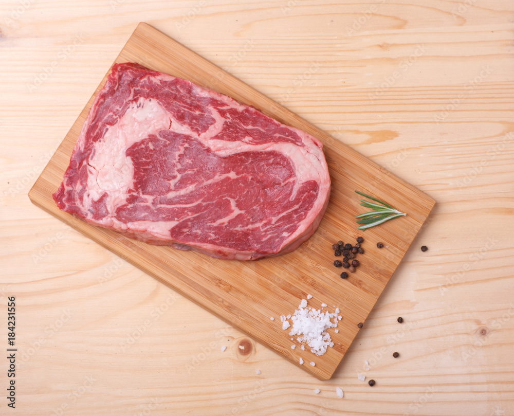 raw uncooked marble beef steak ribeye on cutting board with salt, black pepper, rosemary raw uncooked marble beef steak ribeye on cutting board with salt, black pepper, rosemary