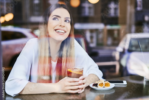 Cheerful young woman having tea and dessert in cafe and looking away