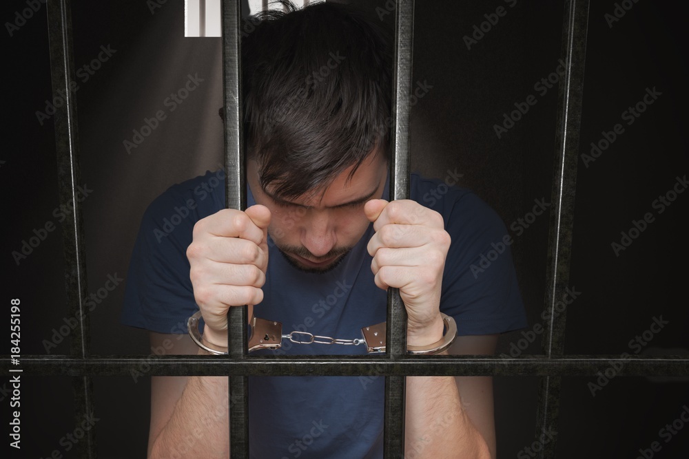 Sad prisoner is holding bars in prison cell. StockFoto Adobe Stock