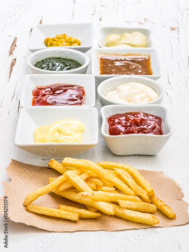 French fries on a piece of crafted paper with different sauces in white sausers on the white wooden background with cracking paint