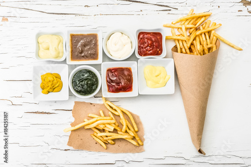 French fries on a piece of crafted paper with different sauces in white sausers on the white wooden background with cracking paint photo