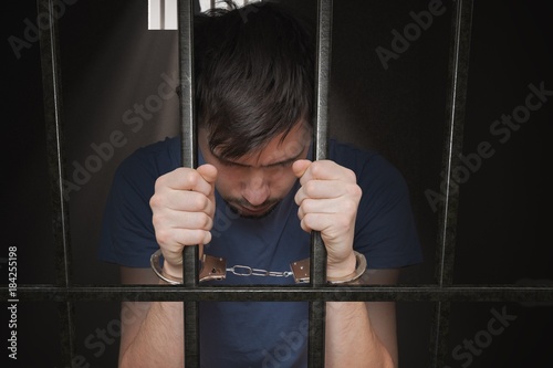 Fototapeta Sad prisoner is holding bars in prison cell.