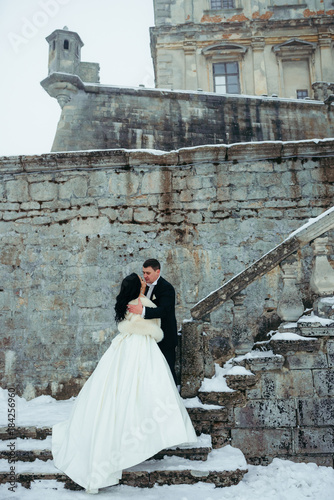 Groom is tenderly hugging the beautiful bride while standing on the snowy stairs of the old house. photo