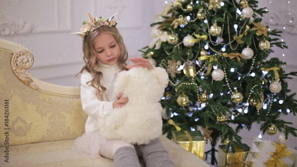 Nice little girl sits on the sofa near the decorated Christmas tree with her soft toy