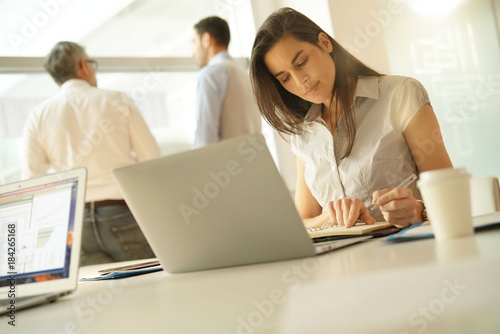 Businesswoman in office working on laptop computer, team in background