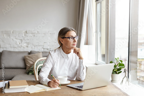 Attractive blonde mature female writer sitting at desk at home placing chin on her hands and looking away with thoughtful or unhappy expression while experiencing writer's block and creative slowdown photo